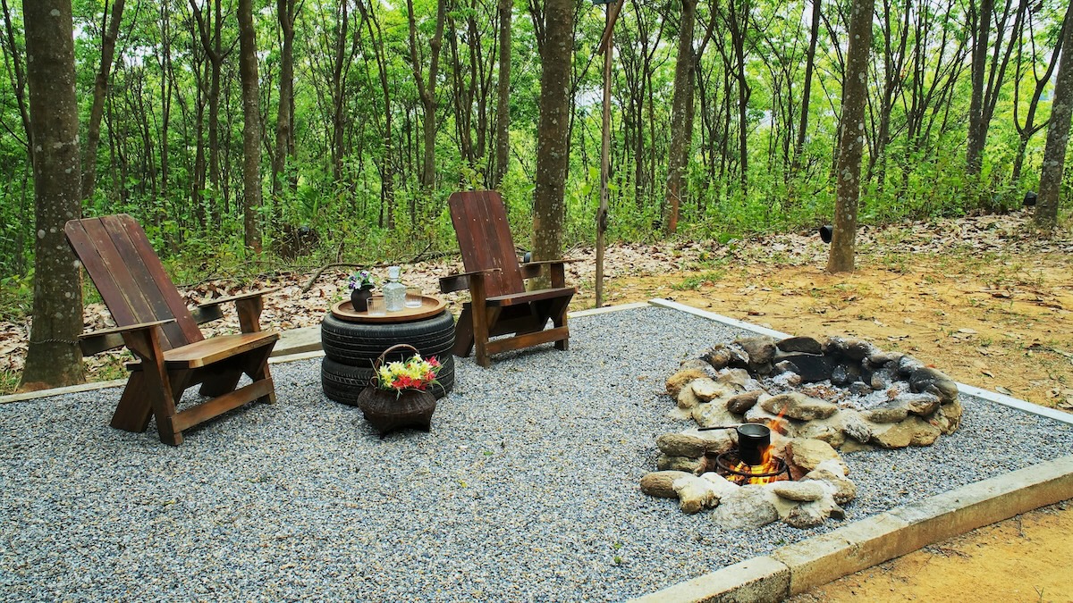 Two rock fire pit with wood table and chairs on gravel floor in the garden background. Stone fire pit with tire table decoration and wooden reclining chair on pebble ground.