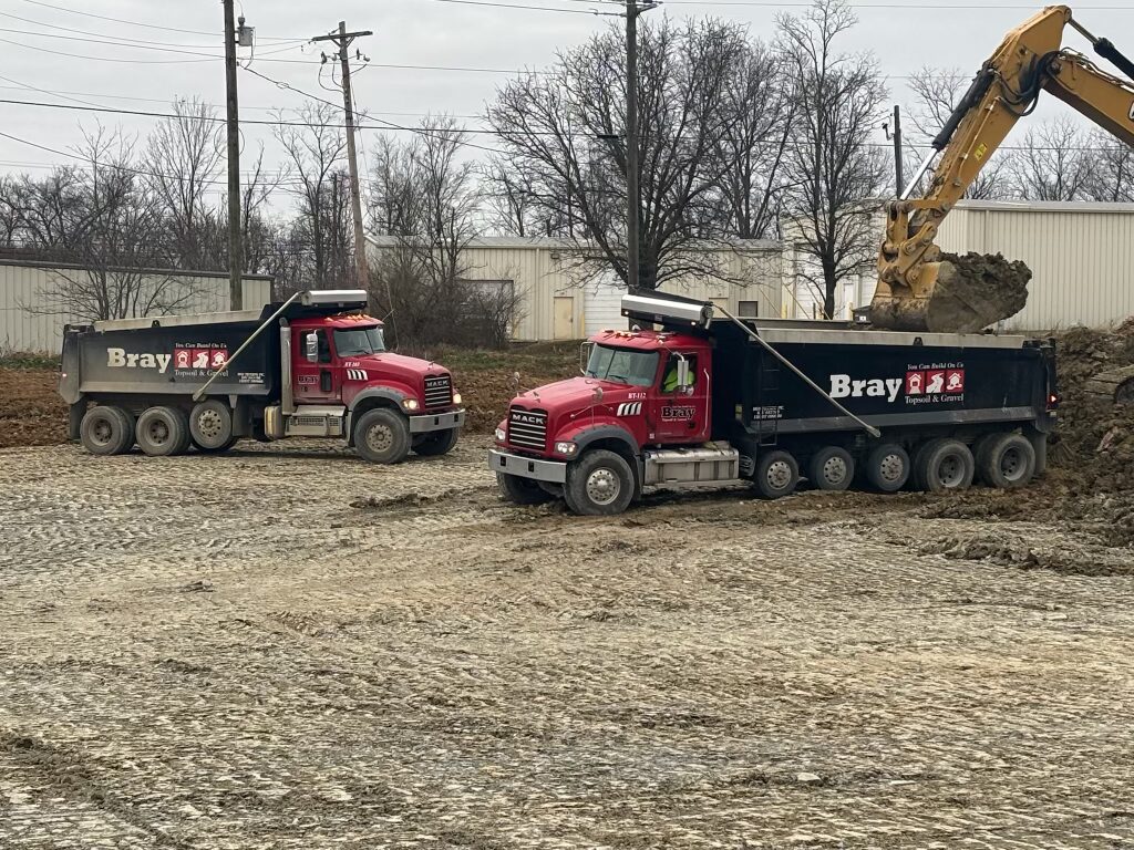 Two red Mack dump trucks at a construction site, one unloading dirt with a yellow excavator in the background.
