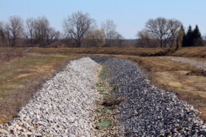 Wide drainage ditch lined with angular riprap stones runs through a cleared field, with sparse grass and bare deciduous trees in the background under a clear blue sky